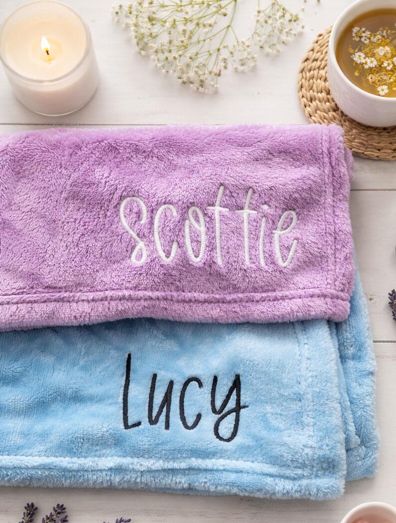 May include: Two plush blankets, one lavender and one light blue, are stacked. The lavender blanket has the name "Scottie" embroidered in white, while the blue blanket has "Lucy" embroidered in black. A lit candle and a cup of tea are in the background.
