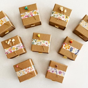 May include: Ten small, square, brown cardboard boxes with drawers. Each box is decorated with a different floral patterned paper tape and a small dried flower.