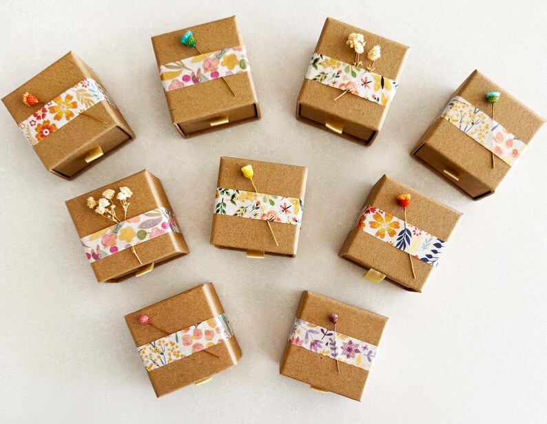 May include: Ten small, square, brown cardboard boxes with drawers. Each box is decorated with a different floral patterned paper tape and a small dried flower.