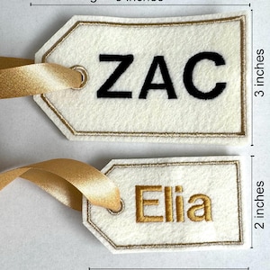 May include: Two personalized gift tags. The larger tag is 5 inches long and 3 inches high, with the name "ZAC" in black. The smaller tag is 3.5 inches long and 2 inches high, with the name "Elia" in gold. Both tags have gold ribbon.
