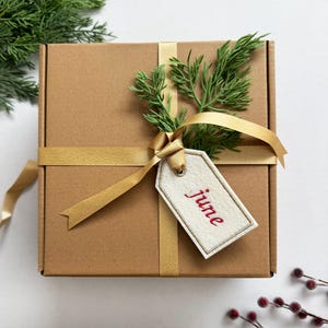 May include: A brown cardboard gift box tied with a gold ribbon and adorned with a sprig of green foliage. A white tag with the red word "june" is attached to the ribbon, suggesting a seasonal or celebratory theme.