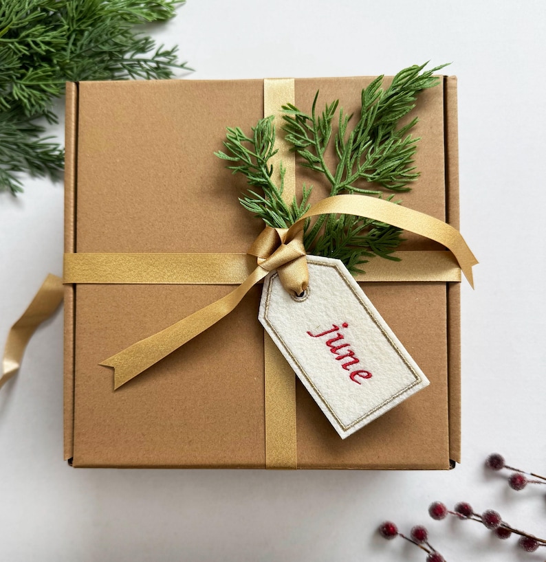 May include: A brown cardboard gift box tied with a gold ribbon and adorned with a sprig of green foliage. A white tag with the red word "june" is attached to the ribbon, suggesting a seasonal or celebratory theme.