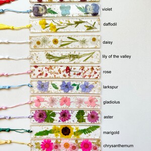 Op de afbeelding: Een set van 12 bladwijzers met geperste bloemen en gouden glitter ingekapseld in helder hars. Elke bladwijzer heeft een andere bloem en is gelabeld met de naam van de bloem: anjer, viooltje, narcis, madeliefje, lelietje-van-dalen, roos, ridderspoor, gladiool, aster, goudsbloem, chrysant en narcis.