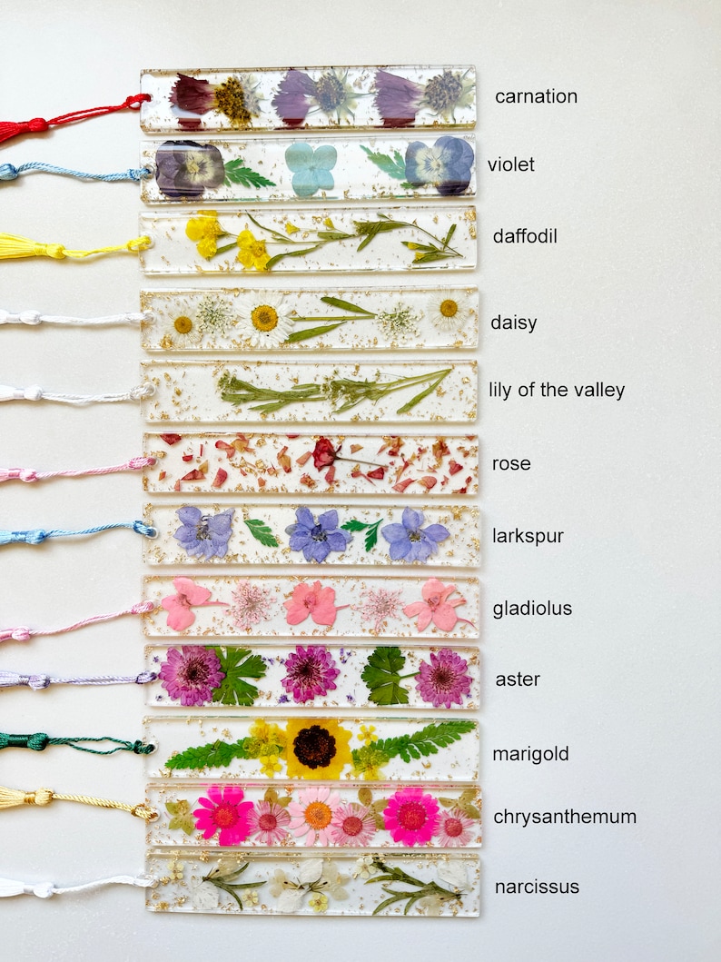 Op de afbeelding: Een set van 12 bladwijzers met geperste bloemen en gouden glitter ingekapseld in helder hars. Elke bladwijzer heeft een andere bloem en is gelabeld met de naam van de bloem: anjer, viooltje, narcis, madeliefje, lelietje-van-dalen, roos, ridderspoor, gladiool, aster, goudsbloem, chrysant en narcis.