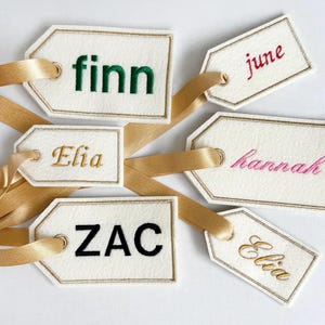 May include: Cream-colored gift tags with gold trim and satin ribbons. Each tag features a name embroidered in a different color: green, gold, red, and black. The names include Finn, Elia, June, Hannah, Zac.