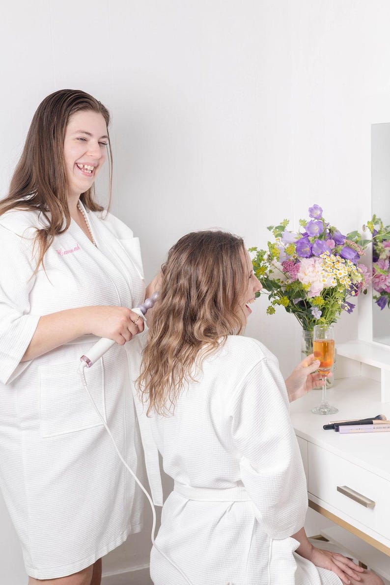 May include: Two women in white robes, one curling the other's hair with a curling iron. A glass of champagne sits on a white vanity with a bouquet of flowers. The scene is bright and cheerful.