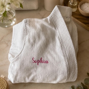 Puede incluir: Una bata de ba&ntilde;o de felpa blanca con un cuello acanalado texturizado y el nombre Sophia bordado en cursiva rosa en la parte delantera izquierda. La bata est&aacute; hecha de una tela suave y absorbente con un cierre frontal plegado y una etiqueta.