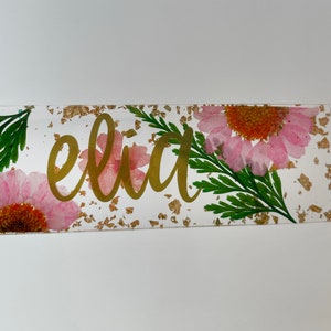 Op de afbeelding: Een transparante rechthoekige bladwijzer met een gouden achtergrond en gouden letters die "eliza" zeggen. De achtergrond is versierd met geperste roze bloemen en bladgoud.