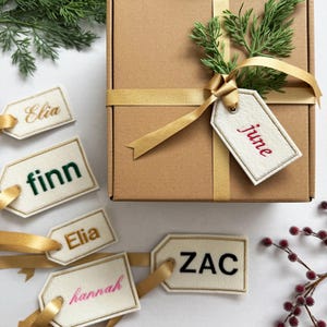 May include: A brown gift box tied with a gold ribbon, adorned with greenery and a tag that says "june" in red. Several name tags with gold ribbon are scattered around the box, each with a different name in various colors.