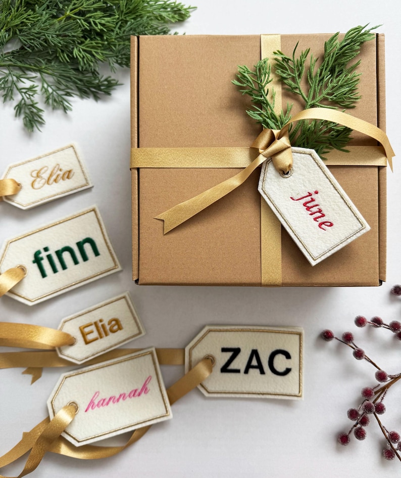 May include: A brown gift box tied with a gold ribbon, adorned with greenery and a tag that says "june" in red. Several name tags with gold ribbon are scattered around the box, each with a different name in various colors.