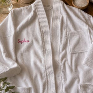 Puede incluir: Una bata de ba&ntilde;o de tela de rizo blanca con cuello acanalado y dos bolsillos delanteros. El nombre Sophia est&aacute; bordado en letra cursiva rosa en el lado izquierdo del pecho. La bata tiene mangas largas y un acabado texturizado de aspecto sencillo.