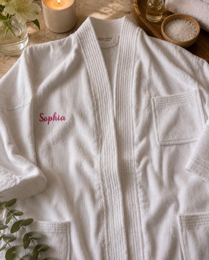 Puede incluir: Una bata de ba&ntilde;o de tela de rizo blanca con cuello acanalado y dos bolsillos delanteros. El nombre Sophia est&aacute; bordado en letra cursiva rosa en el lado izquierdo del pecho. La bata tiene mangas largas y un acabado texturizado de aspecto sencillo.