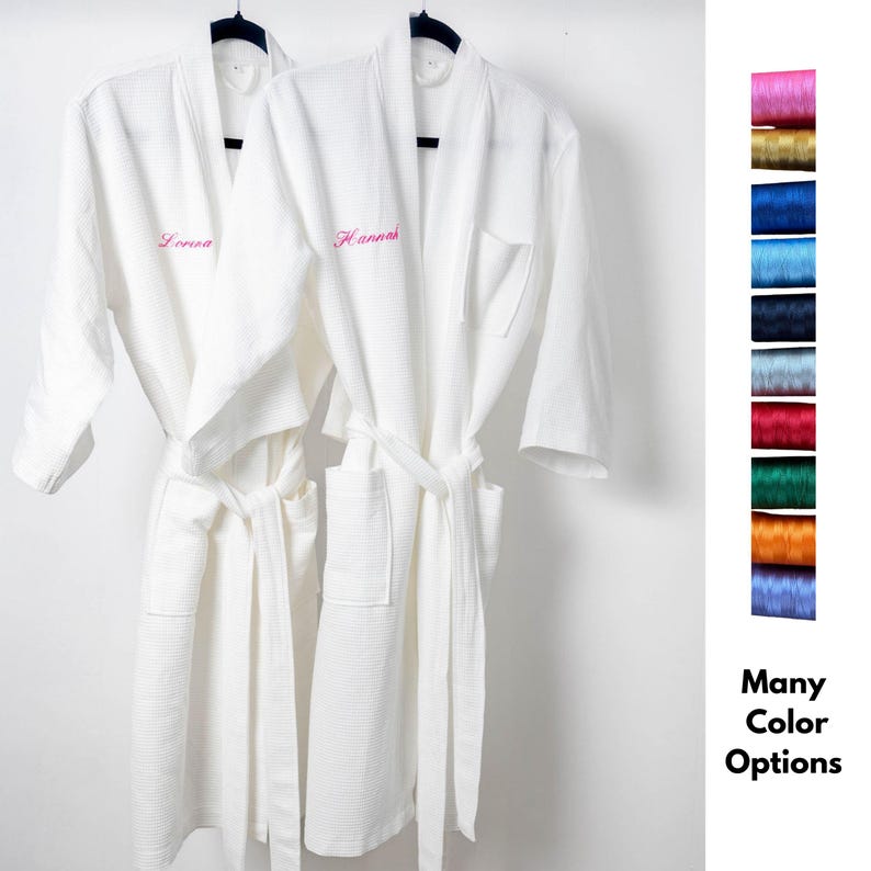 May include: Two white waffle-weave bathrobes hanging on black hangers. Each robe has a matching belt and a pocket. One robe is embroidered with the name "Lorena" in pink, and the other with "Hannah". A color chart shows many thread color options.