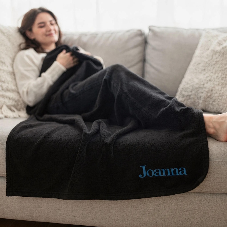 May include: A black plush blanket with the name "Joanna" embroidered in blue. The blanket is draped over a gray sofa. The blanket appears soft and comfortable, perfect for snuggling up.