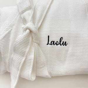 May include: White waffle-weave robe with a tied belt. The robe features the embroidered name "Laolu" in black script on the front. The robe is folded and tied, showcasing the texture and design.