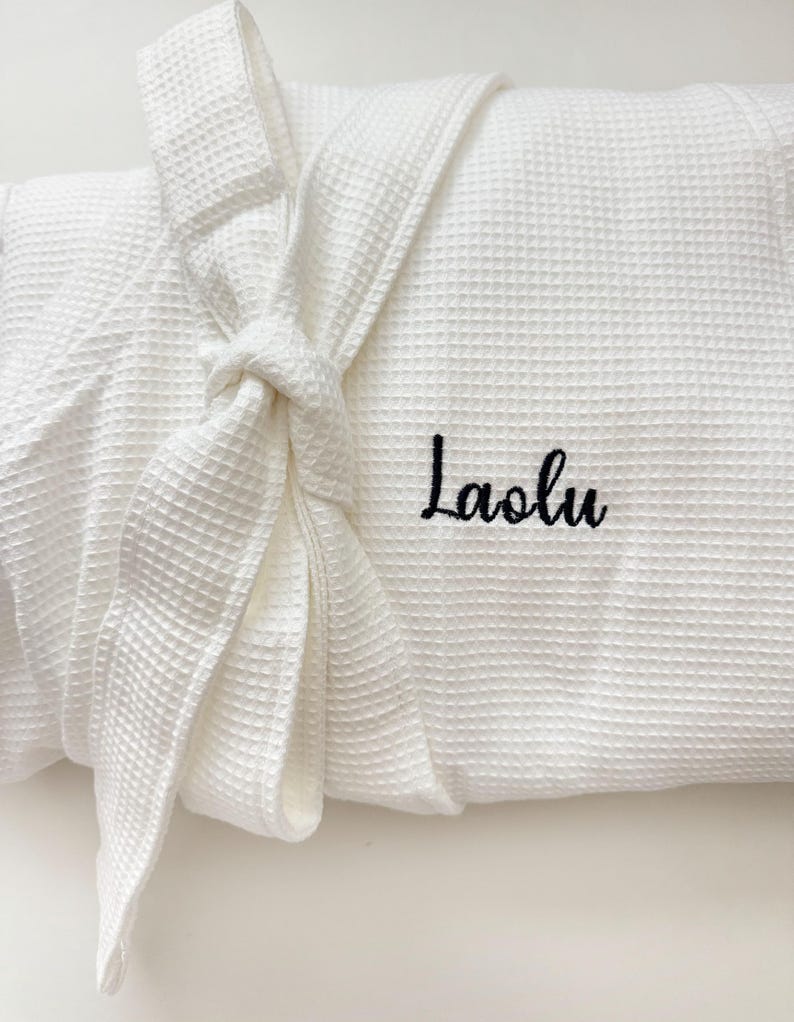 May include: White waffle-weave robe with a tied belt. The robe features the embroidered name "Laolu" in black script on the front. The robe is folded and tied, showcasing the texture and design.