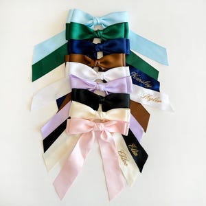 May include: A stack of ten personalized satin hair bows in various colors, including light blue, green, navy blue, brown, white, lavender, black, cream, and pink. Each bow has a gold embroidered name on a white tag.
