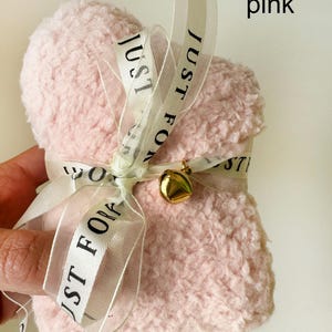 Puede incluir: Una bolsa de coraz&oacute;n de color rosa y peluda con una cinta blanca atada alrededor. La cinta tiene el texto "Just For You" impreso en ella. Una campana dorada est&aacute; unida a la cinta.