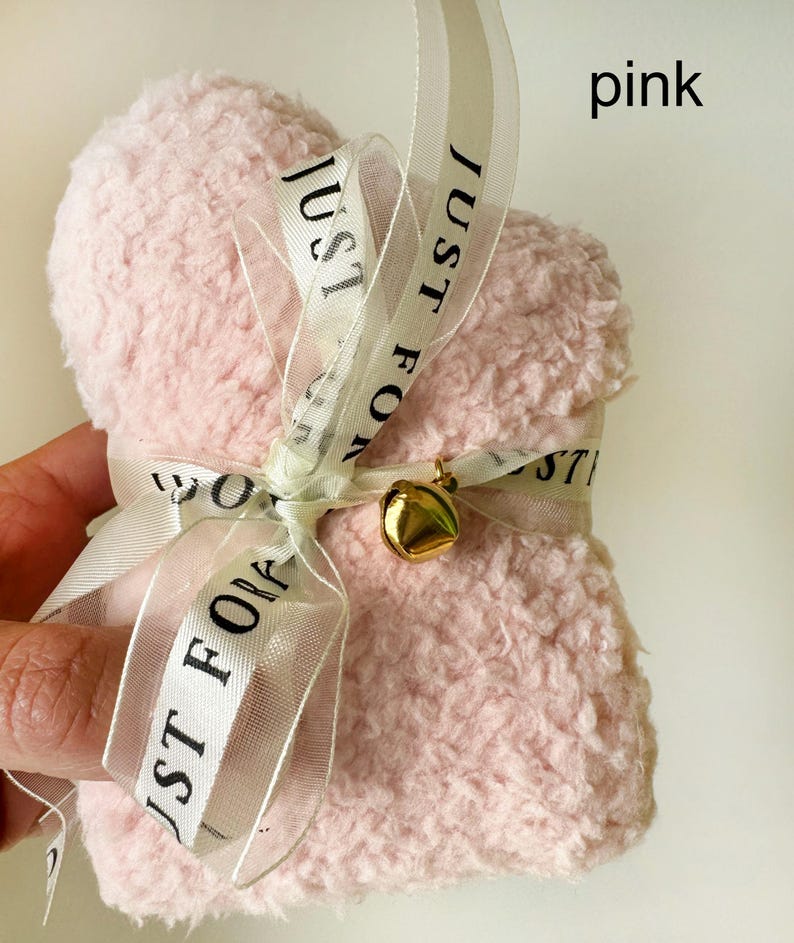Puede incluir: Una bolsa de coraz&oacute;n de color rosa y peluda con una cinta blanca atada alrededor. La cinta tiene el texto "Just For You" impreso en ella. Una campana dorada est&aacute; unida a la cinta.