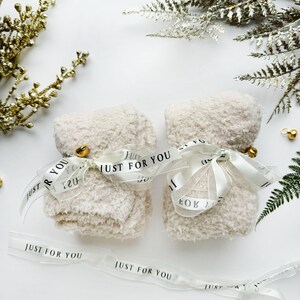Puede incluir: Dos bolsas de regalo blancas y peludas atadas con una cinta transparente con el texto "Just for You" impreso en la cinta. Las bolsas est&aacute;n sobre una superficie blanca con ramas doradas y plateadas de vegetaci&oacute;n.