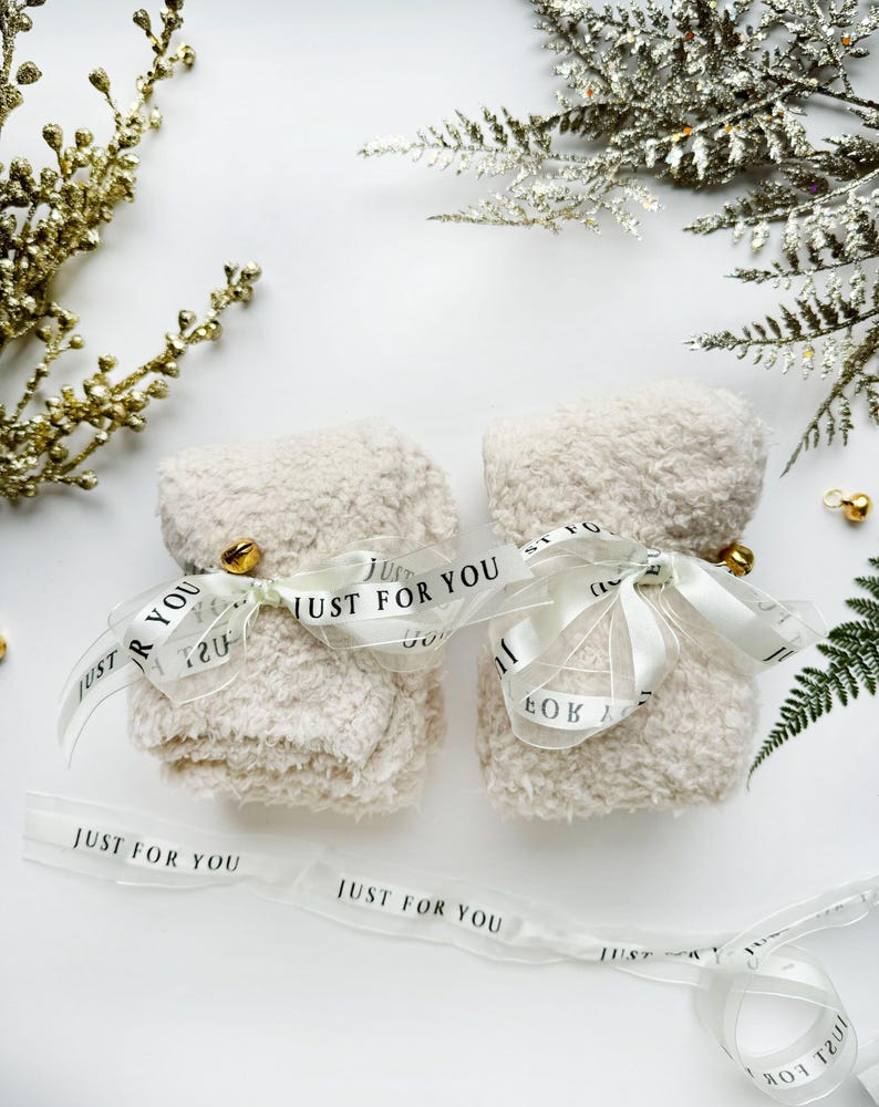 Puede incluir: Dos bolsas de regalo blancas y peludas atadas con una cinta transparente con el texto "Just for You" impreso en la cinta. Las bolsas est&aacute;n sobre una superficie blanca con ramas doradas y plateadas de vegetaci&oacute;n.