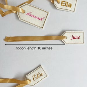 May include: Cream-colored gift tags with gold trim and gold satin ribbons. The tags are embroidered with names in pink and gold thread. The ribbon length is 10 inches.