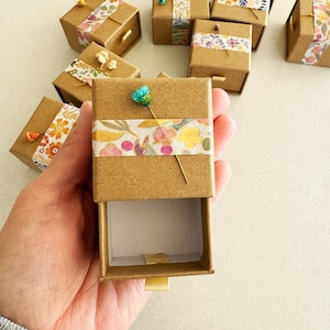May include: A collection of small, brown cardboard boxes with a drawer. Each box is decorated with floral patterned tape and a small flower. The boxes are perfect for storing small items like jewelry or trinkets.