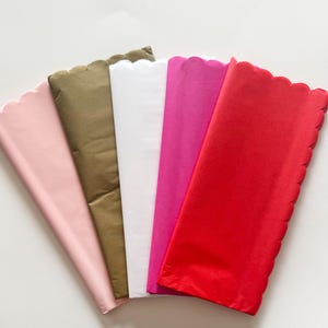 May include: An overhead shot displays a fan of colorful tissue paper sheets. The colors include pink, olive green, white, magenta, and red. Each sheet has a scalloped edge, and they are arranged to show their folded form.