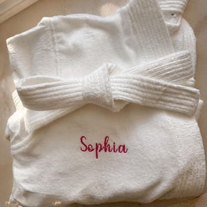 Puede incluir: Una bata de ba&ntilde;o de felpa blanca con un cintur&oacute;n para anudar en la cintura y el nombre Sophia bordado en letras rosas en la parte delantera. La bata tiene un cuello texturizado y una etiqueta de talla M visible cerca del cuello.