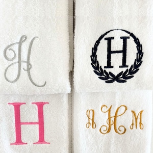 May include: Four white towels with embroidered letter "H" designs. The letters are in silver, navy blue, pink, and gold. The navy blue "H" is encircled by a laurel wreath. The towels are neatly arranged.