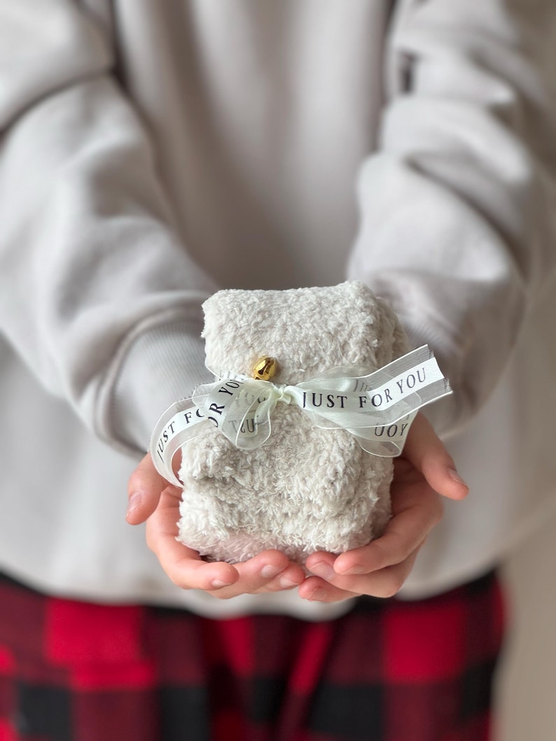 Puede incluir: Un regalo blanco y esponjoso envuelto con una cinta blanca y el texto "Just for you" en negro. El regalo lo sostiene una persona que lleva un su&eacute;ter gris y pantalones de cuadros rojos y negros.