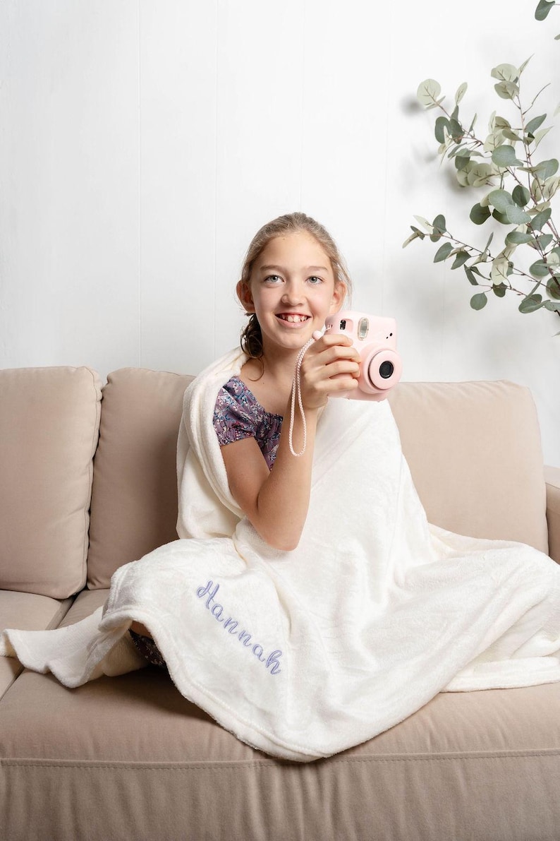 May include: A light pink instant camera with a white strap is held by a person. The person is wrapped in a white blanket with the name "Hannah" embroidered in a light purple script. The person is sitting on a beige couch.