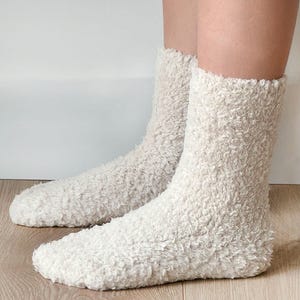 Puede incluir: Un par de calcetines esponjosos de color crema hechos de un material suave y peludo. Los calcetines se muestran en los pies de una persona, con los dedos de los calcetines visibles.