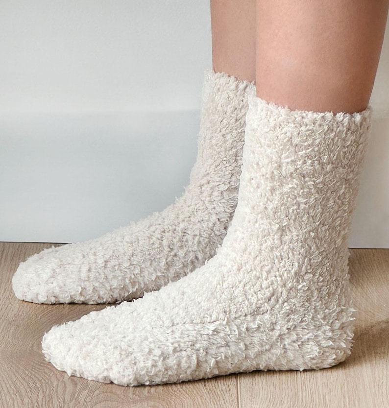 Puede incluir: Un par de calcetines esponjosos de color crema hechos de un material suave y peludo. Los calcetines se muestran en los pies de una persona, con los dedos de los calcetines visibles.