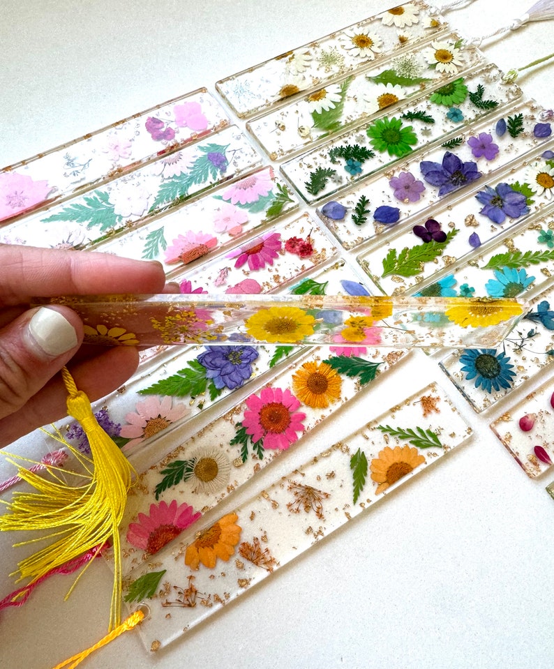 Op de afbeelding: Een verzameling kleurrijke geperste bloemen-bladwijzers met gouden bladaccenten. De bladwijzers zijn gemaakt van helder hars en bevatten een verscheidenheid aan bloemen, waaronder madeliefjes, varens en wilde bloemen. Elke bladwijzer heeft een gele kwast.