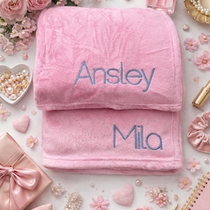 May include: Two pink plush blankets, one folded on top of the other. The top blanket has the name "Ansley" embroidered in blue, and the bottom blanket has "Mila". The blankets are surrounded by pink flowers and decorative items.