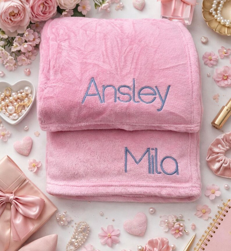 May include: Two pink plush blankets, one folded on top of the other. The top blanket has the name "Ansley" embroidered in blue, and the bottom blanket has "Mila". The blankets are surrounded by pink flowers and decorative items.