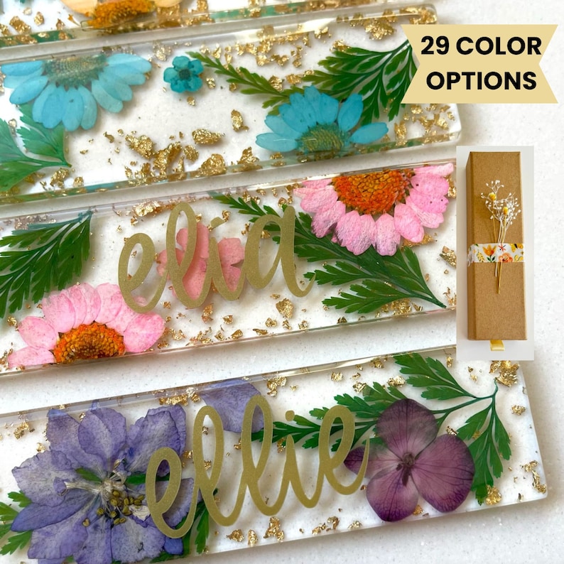 Op de afbeelding: Drie transparante rechthoekige resin boekmerken met geperste bloemen en bladgoud. De boekmerken zijn gepersonaliseerd met de namen "Ellie", "Ella" en "Ellie". De tekst "29 KLEURENOPTIES" is zichtbaar in de rechterbovenhoek van de afbeelding.