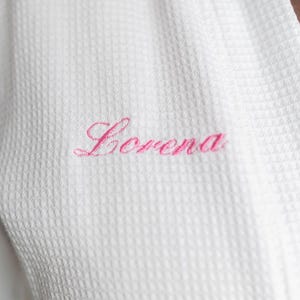 May include: Close-up of a white waffle-weave robe with the name "Lorena" embroidered in pink script. The robe's texture is visible, highlighting the fabric's pattern and the delicate embroidery.