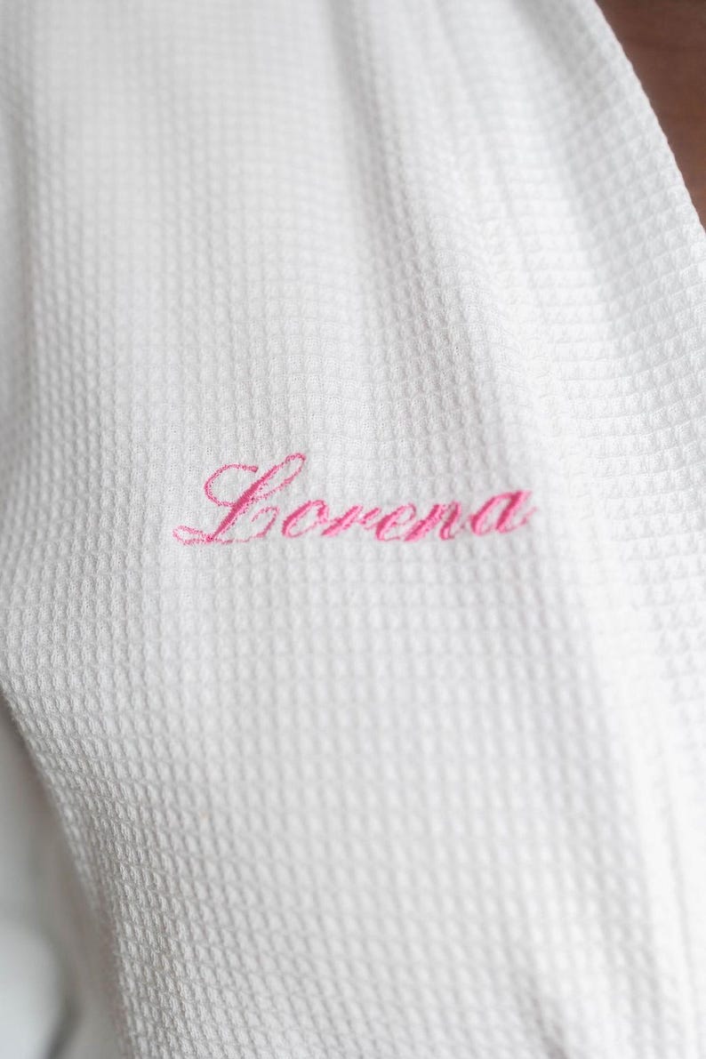 May include: Close-up of a white waffle-weave robe with the name "Lorena" embroidered in pink script. The robe's texture is visible, highlighting the fabric's pattern and the delicate embroidery.