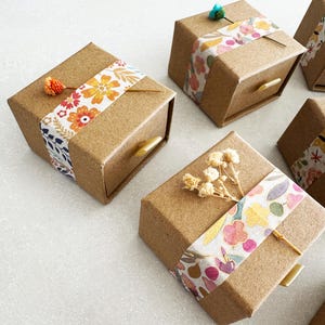 May include: Set of small, brown cardboard boxes with drawers. Each box is decorated with a different floral patterned tape and a small dried flower or sprig.