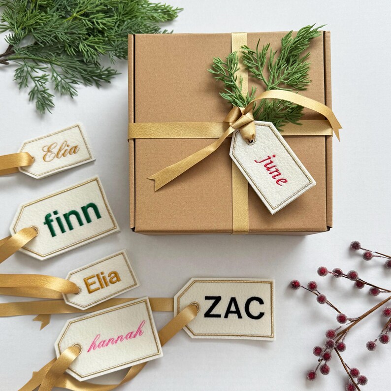 May include: A brown gift box tied with a gold ribbon and adorned with greenery and a tag that says "june". Several personalized gift tags with names like "Elia", "Finn", "Hannah", and "Zac" are attached with gold ribbons.
