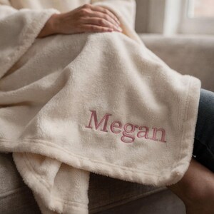 May include: Cream-colored, plush blanket with the name "Megan" embroidered in a soft pink thread. The blanket is folded and draped over someone's lap, suggesting comfort and warmth. The soft texture and personalized detail make it a cozy home accessory.