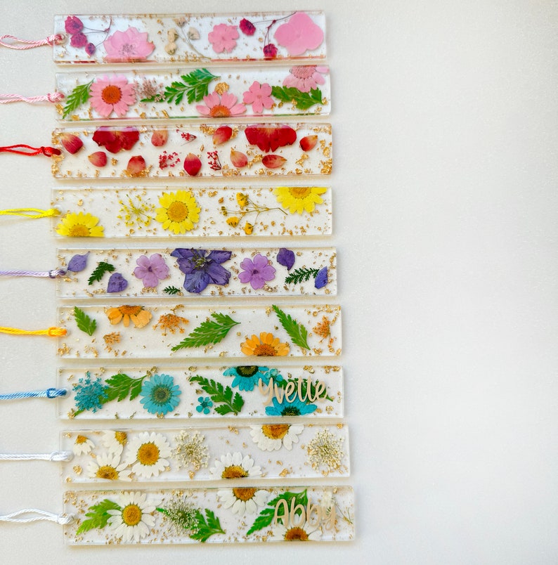 Op de afbeelding: Een set van 10 kleurrijke bladwijzers met geperste bloemen en gouden bladaccenten. Elke bladwijzer heeft een andere bloemenarrangement en een gepersonaliseerde naam.