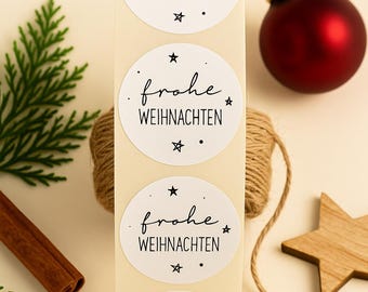 May include: A sheet of white oval stickers with the German phrase "Frohe Weihnachten" (Merry Christmas) in black script. A red Christmas ornament, twine, a wooden star, a cinnamon stick, and greenery are arranged around the stickers.