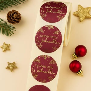 May include: A roll of round Christmas stickers with the text "zauberhafte Weihnachten" in gold script on a burgundy background. The stickers have a gold snowflake and floral design. Christmas ornaments and gold stars are scattered around the roll.