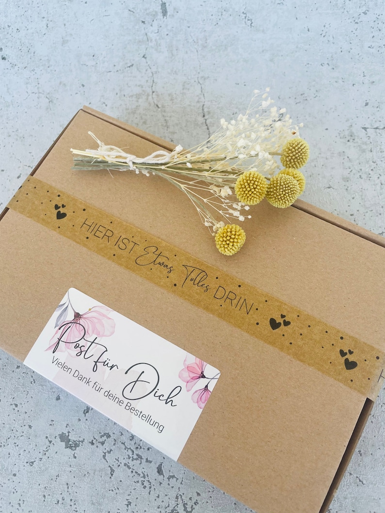 Pu&ograve; includere: Una scatola di cartone marrone con un adesivo bianco che dice "Post f&uuml;r Dich" e "Vielen Dank f&uuml;r deine Bestellung". La scatola &egrave; decorata con un nastro marrone che dice "Hier ist alles drin" e ha piccoli cuori. Un mazzo di fiori secchi bianchi e gialli &egrave; sopra la scatola.