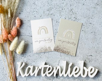 May include: Two cards with rainbow designs and the text "Man ist nie zu klein um großartig zu sein" and "Man ist nie zu klein um großartig zu sein" on a gray background. The cards are next to a white wooden sign that says "Kartenliebe".