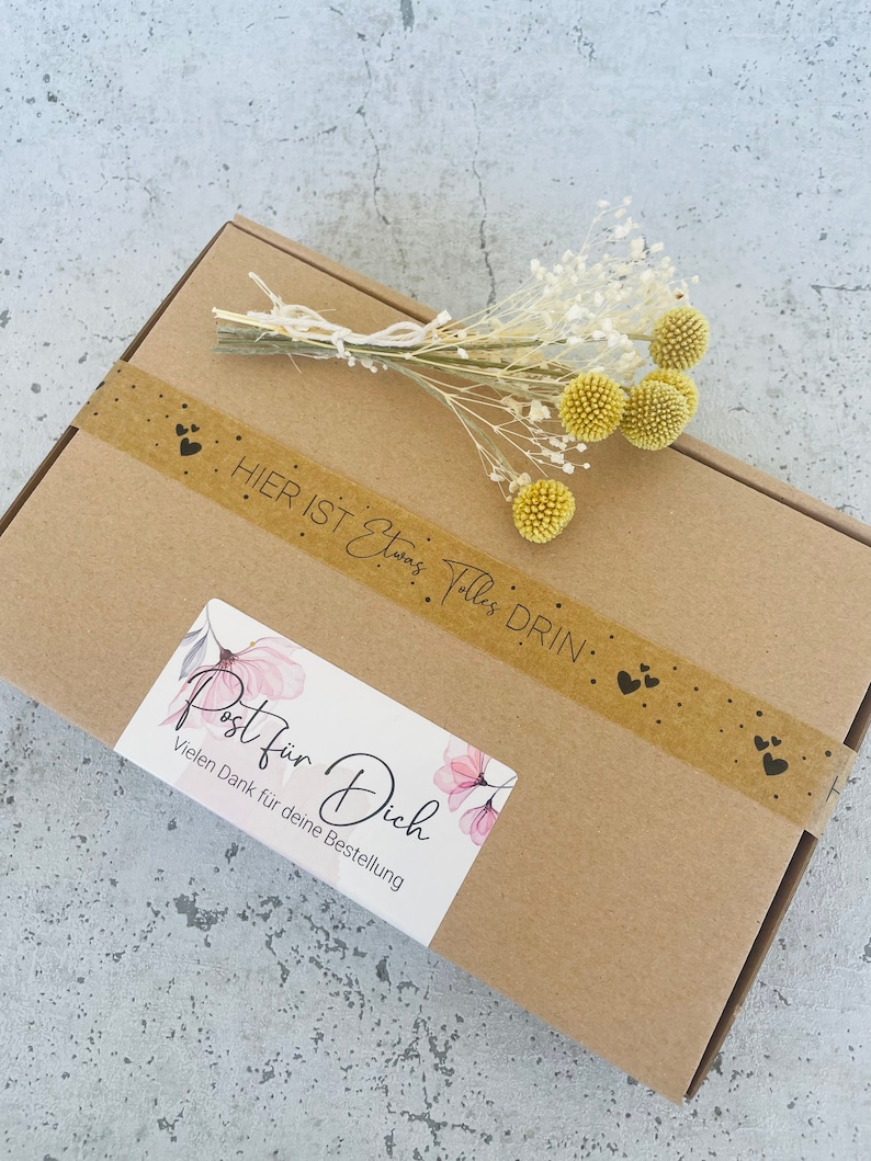 Pu&ograve; includere: Una scatola di cartone marrone con un'etichetta che dice "Post f&uuml;r Dich" e "Vielen Dank f&uuml;r deine Bestellung". La scatola &egrave; decorata con un nastro marrone con cuori e il testo "Hier ist etwas Tolles drin".