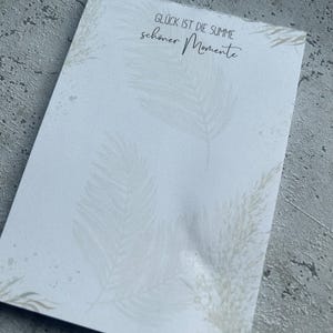 Könnte beinhalten: Weiße Karte mit einem Zitat in schwarzer Schrift: "Glück ist die Summe schöner Momente". Die Karte hat einen Aquarellhintergrund aus weißen Federn.